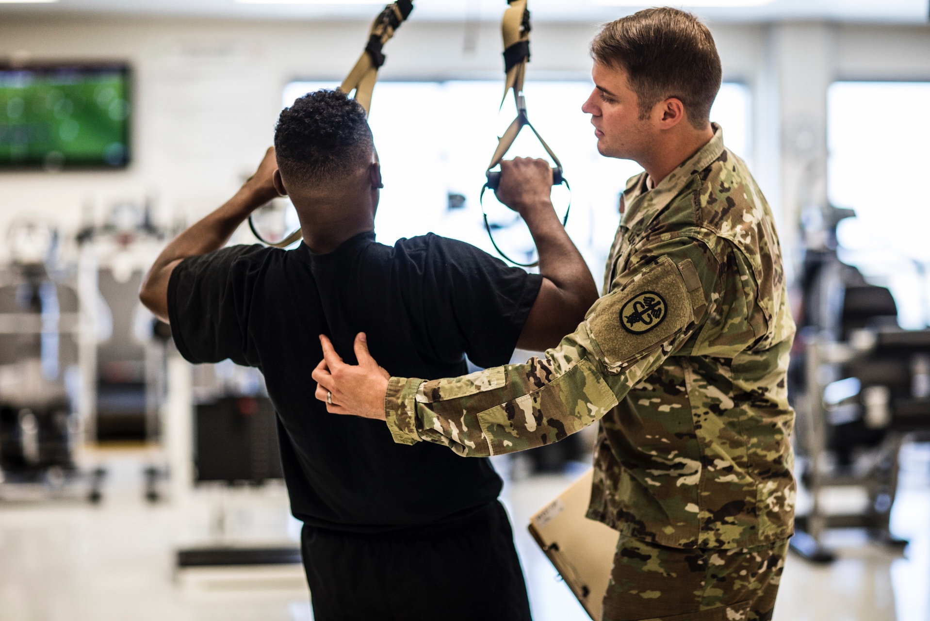 Physical Therapist Staff Sgt. McCoole, Timothy, wearing OCP training with a male soldier wearing APFU inside during the daytime.